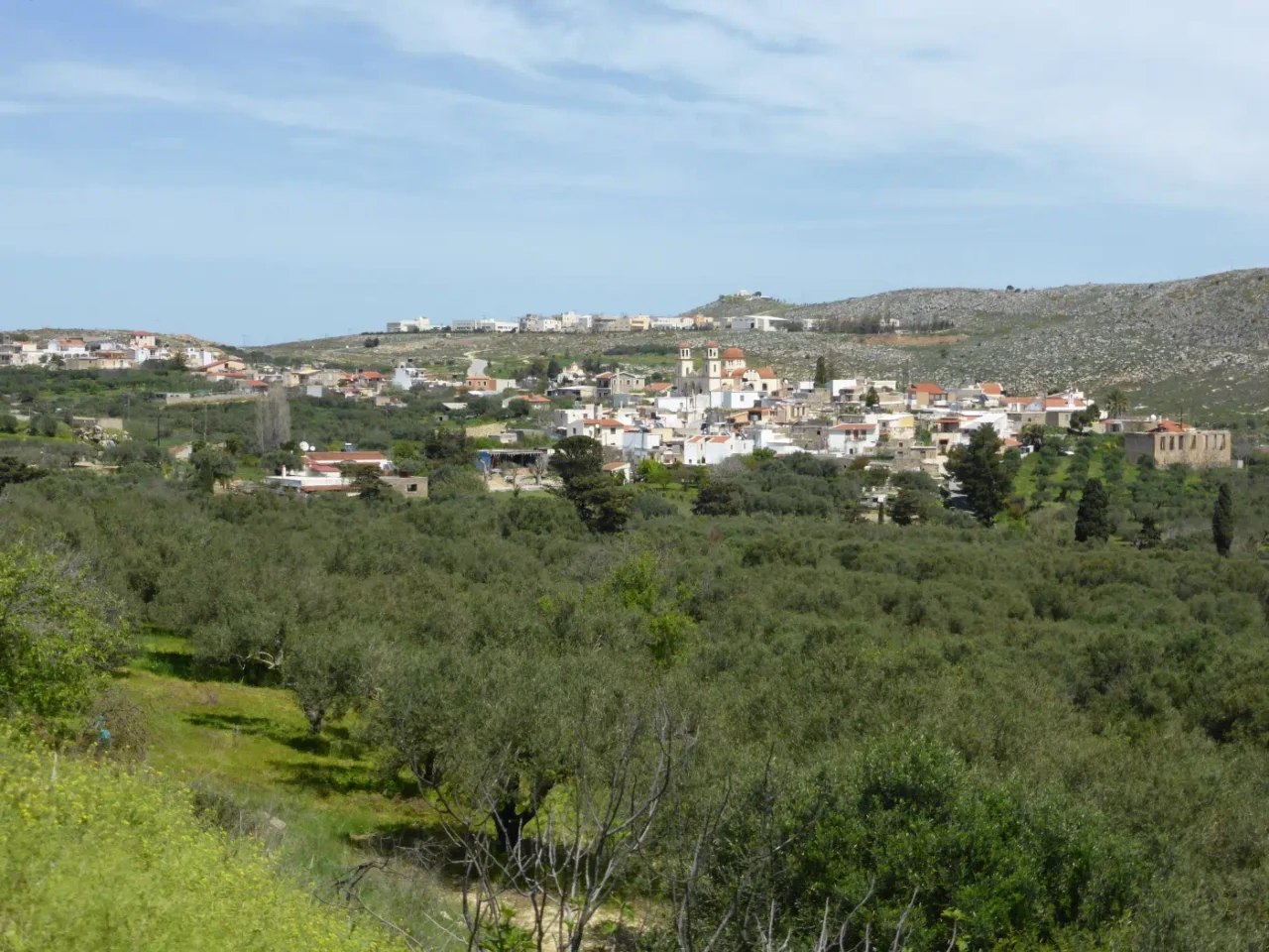 Anopoli view from south-southwest. From right to left, the old school, the old church Taxiarhis, the new church Agia Triada, the cemetery church Panagia, the Handicraft Park in middle ground, Profitis Ilias hilltop chapel in the background. From Wikimedia Commons, Chatev
